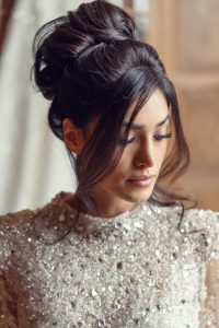 A woman with dark hair styled in an elegant updo wears a sparkling, sequinned dress. She looks down thoughtfully, with soft, natural make-up, against a blurred background.