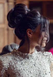 A woman with dark hair styled in an elegant updo and wearing a sparkling, embellished top is seen in profile, with soft lighting and a blurred wooden background.