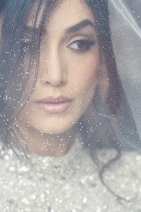A woman with dark hair and subtle make-up gazes thoughtfully to the side, her face partially obscured by a sheer, sparkling veil. She wears a glittering, light-coloured garment.