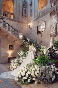 A bride in a white dress with a long train stands on a grand staircase, surrounded by lavish white and green floral arrangements in an ornate hall with statues, tall lamps, and high vaulted ceilings.