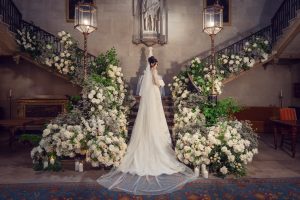 A bride in a long white wedding dress stands between two grand staircases decorated with lush white and green floral arrangements, beneath large lanterns in an elegant, ornate hall.