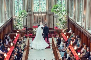 A bride and groom stand at the altar during a wedding ceremony in a grand church, surrounded by guests seated in pews. The church features stained glass windows and large floral arrangements.