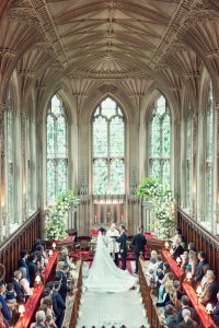 A bride and groom stand at the altar in a grand, ornate chapel with tall arched windows and intricate ceiling details, surrounded by guests seated in wooden pews. Lush floral arrangements decorate the space.