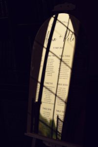A reflective wedding seating plan with “Anna + Peter” and “31 May 2024” at the top, illuminated by sunlight streaming through a window, partially obscuring the list of table allocations.