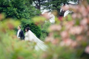 A bride and groom walk outdoors, holding hands, surrounded by lush greenery and blurred pink flowers in the foreground, creating a dreamy, romantic atmosphere.