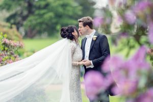 A bride and groom share a romantic kiss outdoors, surrounded by greenery and purple flowers. The bride’s long veil flows behind her as they hold hands, both dressed elegantly for their wedding day.