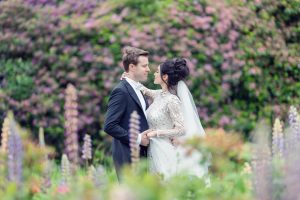 A bride and groom stand close together in a lush garden with blooming flowers and greenery, gazing lovingly into each other's eyes. The bride wears a sparkling gown and veil; the groom is in a black suit.