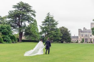 A bride and groom walk hand in hand across a large, green lawn, with the bride’s veil flowing behind her. Tall trees and a grand, castle-like building are visible in the background.