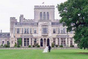 A bride and groom walk hand-in-hand across a green lawn towards a large, historic stone manor with tall windows and an arched entrance. A flag waves atop the building, and a large tree stands to the right.
