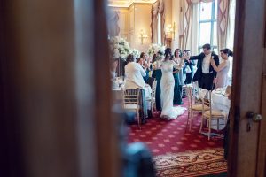 A bride and groom walk hand in hand into an elegant, flower-filled reception room as smiling guests stand and applaud. The photo is taken from behind a partially open door, creating a candid, intimate atmosphere.