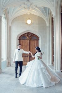 A bride in a voluminous white gown holds a bouquet of white flowers and reaches out to a groom in a white dinner jacket; they stand in an elegant, arched corridor with ornate ceilings and wooden doors.