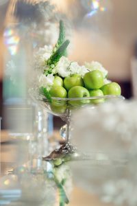 A glass bowl filled with green apples and white flowers sits on a reflective surface, with soft focus and light flares creating a dreamy, elegant atmosphere.