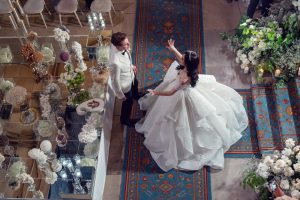 A bride in a voluminous white gown gestures while facing a groom in a white tuxedo jacket. They stand on a patterned blue carpet, surrounded by flowers and elegant decorations, viewed from above.