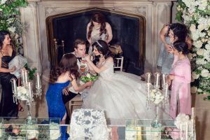 A bride and groom sit together in front of a fireplace, surrounded by women in elegant dresses. The bride wears a white gown and tiara whilst holding a bouquet. The scene features lush white flowers and candles.