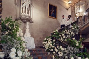 A bride and groom descend an ornate staircase adorned with abundant white flowers and greenery. A multi-tiered wedding cake sits on a table below, and elegant artwork and sculptures decorate the stone walls.