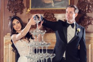 A bride and groom in formal attire pour champagne together into a tall tower of glasses at their wedding, smiling and standing in an elegant, ornate room.