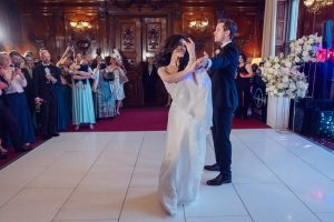A bride and groom dance together on a white dance floor, smiling and holding hands, whilst guests stand around watching and taking photos in an elegant, wood-panelled room decorated with flowers.