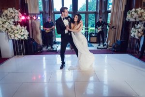 A bride and groom in formal attire share a dance on a white floor, with a live band playing behind them. Large floral arrangements and tall windows with a scenic outdoor view frame the elegant setting.