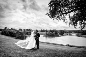 A bride and groom embrace on a grassy hill overlooking a lake and stone bridge, with the bride’s veil flowing in the wind. Trees frame the scenic, cloudy landscape in black and white.