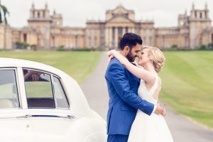 A bride and groom embrace and smile at each other beside a white vintage car, with a grand historic building and spacious grounds in the background.