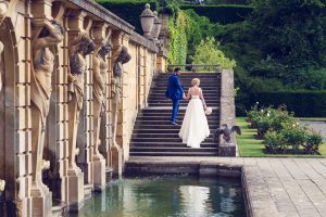 A bride and groom walk up stone steps beside an ornate, classical building with columns and statues, surrounded by lush gardens and a reflecting pool. The bride holds a bouquet and wears a white dress; the groom wears a blue suit.