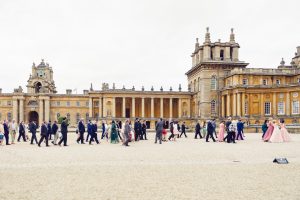 A large group of people, dressed formally, walk across a gravel courtyard in front of an ornate, historic building with columns and towers under an overcast sky.