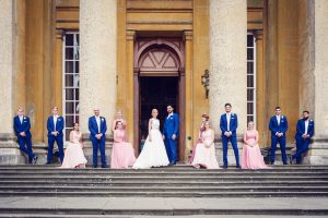 A wedding party stands on stone steps in front of large columns. The bride and groom are in the centre, with bridesmaids in pink dresses and groomsmen in blue suits evenly arranged on either side.