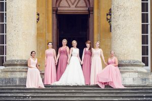 Seven women, including a bride in a white dress and bridesmaids in various shades of pink gowns, pose smiling on stone steps in front of a grand building with large columns and tall doors.