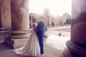 A bride and groom stand close together, gazing into each other's eyes between grand stone columns, with a historic building and soft sunlight in the background.