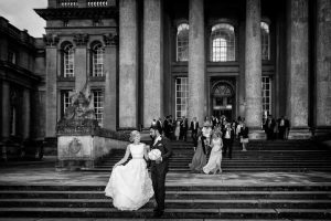 A bride and groom walk down the steps of a grand building with tall columns, smiling at each other, whilst wedding guests follow behind them. The setting is elegant and formal, captured in black and white.