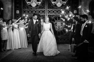 A bride and groom walk hand in hand, smiling, whilst guests on either side hold sparklers in celebration. The scene is at night in front of ornate gates. The photo is in black and white.