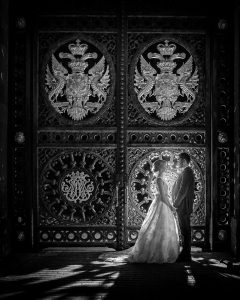 A bride and groom stand facing each other, holding hands in front of an ornate, large gate decorated with intricate patterns and double-headed eagle emblems, with sunlight casting dramatic shadows.