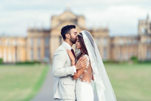 A groom in a light suit gently kisses the forehead of his bride, who wears a white lace wedding dress and veil. They embrace outside in front of a large, elegant building with blurred architecture.