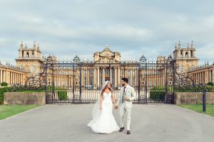 A bride and groom walk hand in hand, smiling, in front of an ornate iron gate and a grand historic building with columns and towers under a cloudy sky.
