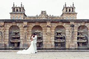A bride and groom embrace in front of an ornate stone wall with statues and fountains, with a grand historic building in the background. The bride wears a white dress and the groom a light suit.
