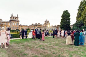 A large group of people dressed in formal and colourful attire gathers on the lawn of a grand historic building with ornate architecture and gardens, socialising at an outdoor event.