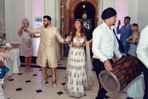 A woman in a white and gold lehenga and a man in a cream sherwani smile and hold hands whilst dancing at a celebration; a drummer leads the group, and other guests watch and join in.