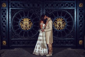 A couple in elaborate traditional South Asian attire stands closely together, touching foreheads, in front of ornate black and gold gates with intricate circular and floral designs.