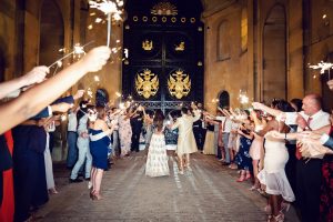 A couple walks hand in hand through a tunnel of guests holding sparklers at night, celebrating outside a grand gate adorned with gold crests. The festive crowd lines both sides, clapping and cheering.