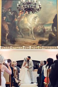 A bride and groom share a kiss at their indoor wedding ceremony, surrounded by seated guests. A large, dramatic painting and chandelier hang above them, creating a grand and elegant atmosphere.
