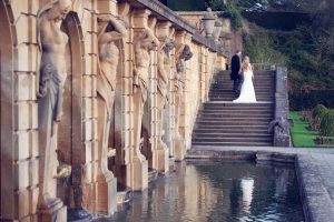 A bride and groom stand on stone steps beside a reflective pool and ornate columns with carved statues in a formal garden setting. The bride wears a white dress, and the groom wears a black suit.