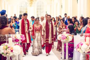 A bride in a white and red lehenga, escorted by two men in traditional attire, walks down an aisle lined with guests and floral arrangements at a vibrant Indian wedding ceremony.