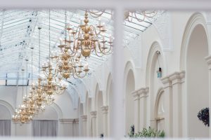 Elegant chandeliers hang from a high arched glass ceiling in a grand, white hall with intricate detailing and large windows. Ornate columns and lush green plants decorate the sophisticated interior.