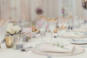 Elegant table setting with crystal glasses, gold accents, white plates, and floral centrepieces featuring white and pink flowers. The soft, blurred background suggests a formal event or wedding reception.
