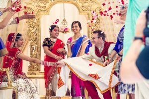 A joyful Hindu wedding ceremony with the bride and groom smiling and holding hands, surrounded by colourful, traditionally dressed guests, flower petals in the air, and ornate golden decor in the background.