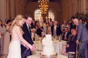 A bride and groom, smiling, cut their wedding cake together in a grand hall whilst guests watch and celebrate around them. The cake is white with flowers, and a chandelier hangs above.