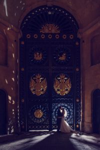A bride and groom stand embracing in front of an ornate, tall, black and gold gate at night, with soft light illuminating them and intricate patterns visible on the gate.