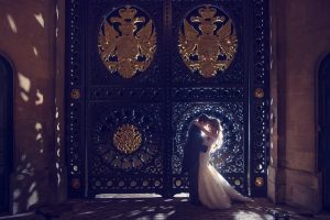 A bride and groom embrace and kiss in front of a large, ornate wrought iron gate decorated with gold designs and illuminated by soft, dramatic lighting.