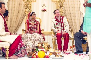 A bride and groom in traditional Indian wedding attire sit on ornate gold chairs during a wedding ceremony, surrounded by guests and decorative elements, with ceremonial items arranged on the floor in front of them.