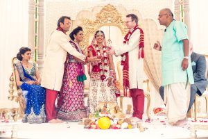 A joyful South Asian wedding ceremony shows the bride and groom in traditional attire, flanked by family members, exchanging ritual items and smiling, with ornate golden decor in the background.